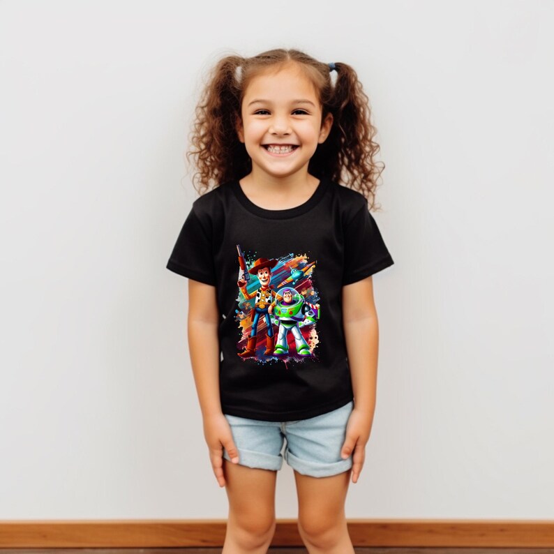 May include: A child wearing a black t-shirt with a vibrant graphic of Woody and Buzz Lightyear from Toy Story. The shirt is worn with light blue denim shorts. The background is a plain white wall.