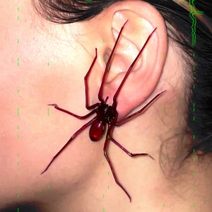 May include: A red plastic spider used as an earring. The spider is large and has long legs. The text "Plastic Spider as an Earring" and "[Ruby Red]" is visible in the image.