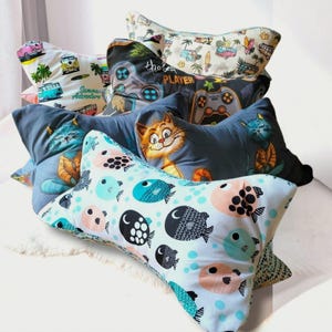 Könnte beinhalten: Eine Sammlung knochenförmiger Kissen mit verschiedenen Mustern, darunter ein hellblaues Kissen mit schwarzen, rosa und türkisfarbenen Fischen, ein dunkelgraues Kissen mit einer Comic-Katze, ein schwarzes Kissen mit Gamecontroller und ein cremefarbenes Kissen mit Vans.