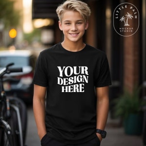 Könnte beinhalten: Ein schwarzes T-Shirt mit dem Text "YOUR DESIGN HERE" in weißen Buchstaben. Das Shirt wird von einem jungen Menschen getragen, der in einer städtischen Umgebung steht.