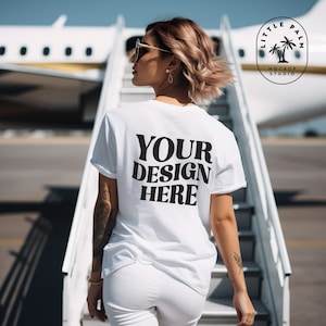 May include: A white t-shirt with the text "YOUR DESIGN HERE" printed on the back. The t-shirt is being worn by a person standing on a set of stairs in front of an airplane.