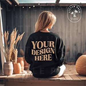 May include: A black sweatshirt with the text "YOUR DESIGN HERE" in a tan color. The sweatshirt is being worn by a person sitting on a wooden bench.