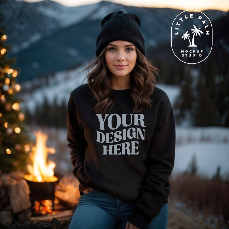 May include: A black sweatshirt with the text "YOUR DESIGN HERE" in white. The sweatshirt is being worn by a person with long brown hair and a black beanie. The person is standing in front of a fire and a snowy mountain background.