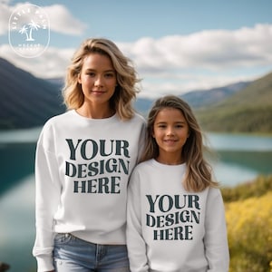 Könnte beinhalten: Zwei weiße Crewneck-Sweatshirts mit dem Text "YOUR DESIGN HERE" auf der Vorderseite. Die Sweatshirts werden von einer Frau und einem Mädchen getragen, die vor einem See und Bergen stehen.