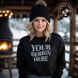 May include: A black sweatshirt with the text "YOUR DESIGN HERE" in white. The sweatshirt is being worn by a person with blonde hair and a black beanie with a brown pom-pom.