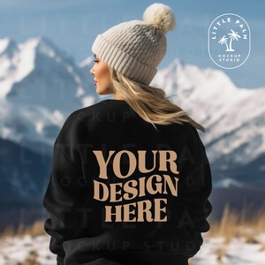 May include: A black sweatshirt with the text "YOUR DESIGN HERE" in brown lettering. The sweatshirt is being worn by a person with blonde hair and a white beanie. The person is standing in front of a snowy mountain background.