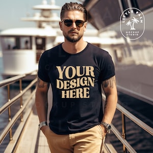 Könnte beinhalten: Ein Mann trägt ein schwarzes T-Shirt mit dem Text "YOUR DESIGN HERE" in brauner Schrift. Er steht auf einem Steg mit einem großen weißen Boot im Hintergrund.