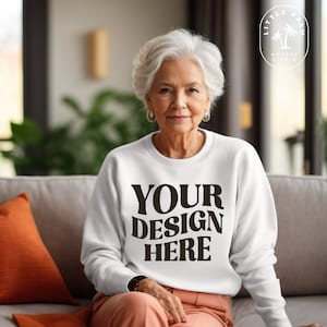 May include: A white sweatshirt with the text "YOUR DESIGN HERE" printed in brown on the front. The sweatshirt is being worn by a person sitting on a couch.