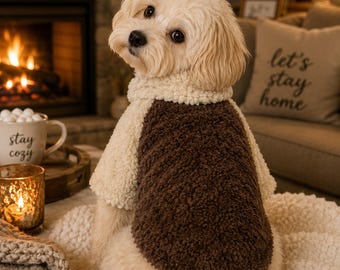 May include: A small, light-colored dog with floppy ears wearing a two-toned fleece sweater. The sweater features a cream-colored collar and sleeves with a dark brown, textured fleece panel covering the back, providing a soft, fuzzy appearance.
