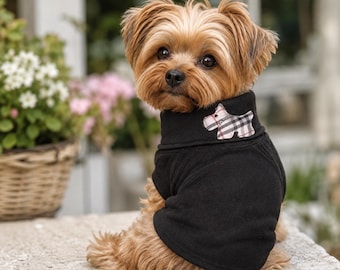 Puede incluir: Un perro pequeño de color canela con pelaje largo y suave que lleva un suéter negro de cuello alto. El suéter presenta un pequeño parche bordado en la zona del cuello con forma de perro, decorado con un patrón de cuadros beige, negro y rojo.