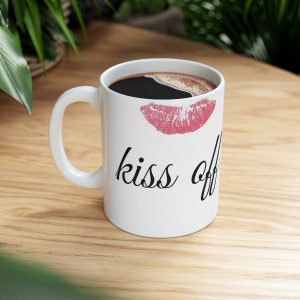 Könnte beinhalten: Weiße Keramik-Kaffeetasse mit einem rosafarbenen Lippenstift-Abdruck und dem Text "kiss off" in schwarzer Kursivschrift.