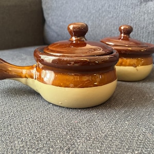 May include: Two ceramic, lidded soup bowls. The bowls are brown with a cream-colored base. One bowl has a handle. The lids have a small knob on top. The bowls are on a gray surface.