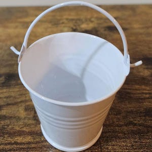 May include: A small white metal bucket with a handle. The bucket is empty and has a slightly textured surface.