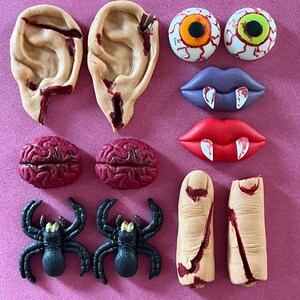 Puede incluir: Un conjunto de decoraciones de fondant con temática de Halloween, que incluyen orejas, ojos, labios, cerebros, arañas y dedos. Las decoraciones están hechas de fondant y están pintadas con detalles rojos y negros.