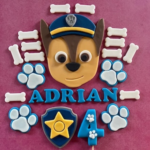 Op de afbeelding: Taartdecoratie met een cartoonhondengezicht met een blauwe politiepet, omringd door botvormige snoepjes en pootafdrukken. De naam "ADRIAN" staat in blauwe letters, samen met het nummer "4" en een badge.