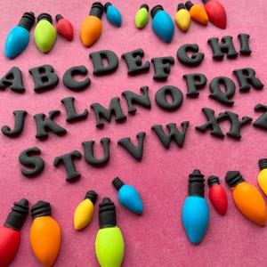 Op de afbeelding: Een verzameling kleurrijke, decoratieve gloeilampen en zwarte alfabetletters op een roze achtergrond. De gloeilampen zijn in verschillende kleuren, waaronder rood, oranje, geel, groen en blauw. De letters zijn vetgedrukt en schreefloos.