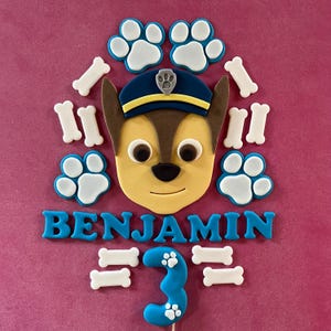 May include: A cake decoration with a cartoon character's face, blue paw prints, and bone shapes. The name "BENJAMIN" is in blue letters, with a blue number "3" on a stick. The background is a solid pink colour.