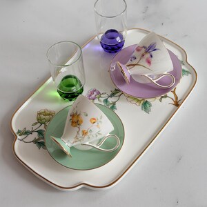 Puede incluir: Una bandeja blanca con borde dorado, que contiene dos tazas de té con platillos y dos vasos pequeños. Las tazas y los platillos son de colores pastel con diseños florales. Los vasos tienen bases de colores, uno verde y otro morado.