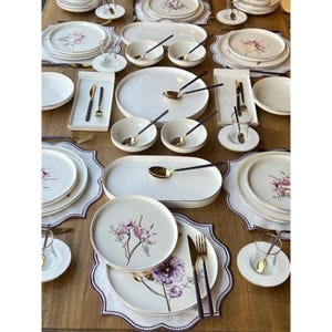May include: A dinnerware set featuring white ceramic plates, bowls, and rectangular platters with delicate purple floral illustrations and gold-toned rims. The collection includes matching glasses, forks, and knives with dark handles arranged on placemats.