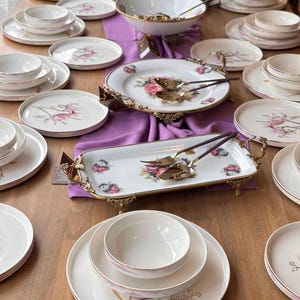 May include: A collection of white porcelain dinnerware featuring floral patterns and gold-toned rims. The set includes round plates, bowls, and rectangular serving trays with ornate gold metal handles and feet, accompanied by gold-toned cutlery.