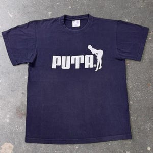 May include: A dark navy blue short-sleeved t-shirt featuring a white graphic print on the chest. The graphic displays the word PUTA in bold capital letters, with a white silhouette of a person leaning over positioned to the right of the text.