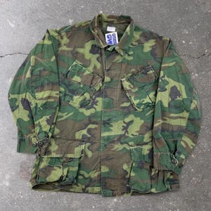 Puede incluir: Una chaqueta de manga larga con botones y estampado de camuflaje boscoso en tonos verde oscuro, verde claro, marrón y negro. La prenda incluye dos bolsillos en el pecho con solapa y dos bolsillos delanteros inferiores con solapa, además de una etiqueta blanca.