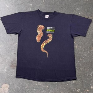 May include: A black short-sleeve t-shirt featuring two brown and yellow patterned snakes printed on the front. The text reads Anniston Museum of Natural History REPTILES The Beautiful and the Deadly, with dates September 14, 2009-January 10, 2010.