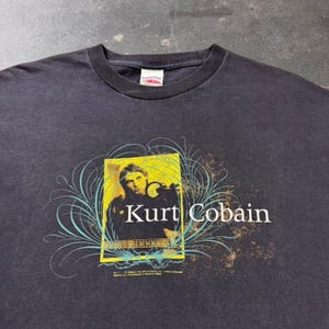 Puede incluir: Una camiseta negra de cuello redondo con un gráfico rectangular amarillo de Kurt Cobain tocando la guitarra. El diseño incluye el texto blanco Kurt Cobain y líneas curvas de color azul claro que rodean la imagen central.
