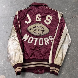 May include: A maroon satin varsity jacket with cream-colored sleeves. The back features cream-colored lettering that reads J & S MOTORS, with a central oval patch displaying MEMPHIS 47-48-49-50 CHAMPS. It has a ribbed collar, cuffs, and hem.