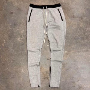 Op de afbeelding: Grijze joggingbroek met een zwarte tailleband en zwarte ritsen op de zakken en enkels. De broek heeft een trekkoord en een relaxte pasvorm. Het materiaal lijkt een zachte, comfortabele stof te zijn.