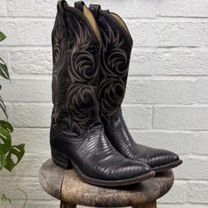 Könnte beinhalten: Ein Paar schwarze Leder-Cowboystiefel mit einer strukturierten Eidechsenhaut-Optik am Fußteil und einem hohen Schaft, der mit aufwendigen, geschwungenen Ziernähten in Dunkelbraun versehen ist, inklusive Zuglaschen an der Oberseite jedes Stiefels.