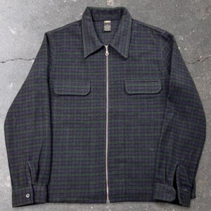 May include: A long-sleeve jacket featuring a dark green and navy blue plaid pattern. This garment includes a full-length center front zipper, a pointed collar, two chest pockets with flaps, and buttoned cuffs on each sleeve. A small label is visible.