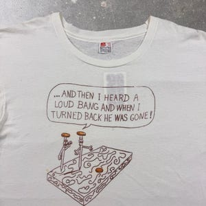以下が含まれることがあります： 白いTシャツに、質感のある表面に立つ2本の擬人化された釘の茶色の線画が描かれています。その上の吹き出しには「... AND THEN I HEARD A LOUD BANG AND WHEN I TURNED BACK HE WAS GONE!」というテキストが含まれています。