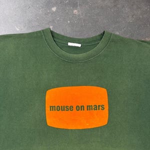 May include: A dark olive green crew neck t-shirt featuring a rectangular orange graphic patch centred on the chest. The text "mouse on mars" is printed in dark green lowercase letters across the centre of the orange rectangular graphic.