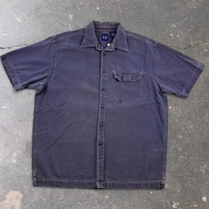 Puede incluir: Una camisa de manga corta con botones en un color gris carbón deslavado. La camisa presenta un cuello estándar, un bolsillo en el pecho con solapa abotonada y una tapeta central con botones. La etiqueta interior muestra la marca GAP.