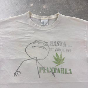 Op de afbeelding: Een wit T-shirt met korte mouwen en een zwarte lijntekening van een stokfiguur met een sceptische uitdrukking. De opdruk bevat de groene tekst BASTA... E ORA DI PIANTARLA en een illustratie van een groen cannabisblad op de voorzijde.