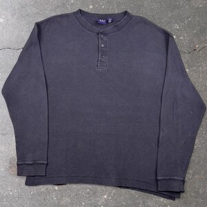 Könnte beinhalten: Ein dunkel anthrazitfarbenes langärmeliges Henley-Shirt mit Waffelstrick-Textur, Rundhalsausschnitt und einer Knopfleiste mit drei Knöpfen. Das Etikett am Innenkragen zeigt den Markennamen GAP in weißer Schrift auf blauem Grund.