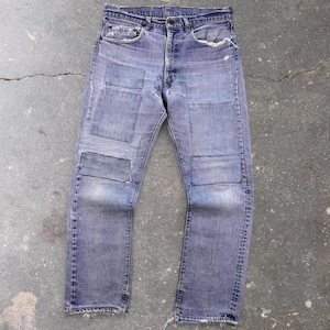 Può includere: Un paio di jeans in denim azzurro con finitura usurata e toppe rettangolari visibili sulla parte anteriore di entrambe le cosce e le ginocchia. I jeans presentano una chiusura a bottoni, cinque tasche e bordi sfilacciati in vita e sul fondo.