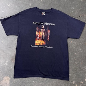 May include: A dark navy blue short-sleeved t-shirt featuring a central graphic print of a skeleton display. The text above the image reads Mütter Museum, and the text below reads The College of Physicians of Philadelphia in white lettering.