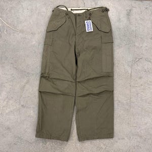 Puede incluir: Pantalones cargo verde oliva con múltiples bolsillos y cierre de botón. Los pantalones tienen un ajuste relajado y una etiqueta que dice "DUAG". Estos pantalones son un estilo clásico de inspiración militar.