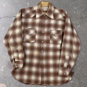 Peut inclure: Une chemise à manches longues boutonnée avec un motif à carreaux marron et crème. La chemise comprend deux poches de poitrine à rabats pointus, un col pointu et une doublure de col beige. L'avant est fermé par une rangée verticale de boutons.