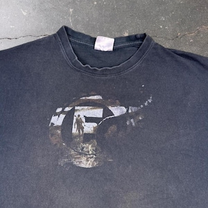 Puede incluir: Una camiseta negra de cuello redondo con un gran gráfico circular en el pecho. El gráfico contiene una imagen desgastada en blanco y gris de una persona de pie en una puerta, enmarcada por una letra F en negrita. La prenda tiene aspecto usado.