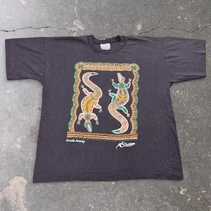 May include: A black short-sleeve t-shirt featuring a central graphic of two stylized crocodiles within a decorative rectangular border. The text "Crocodile Dreaming" and "Churinga" is printed below the graphic in white, with orange and green accents.