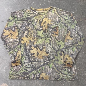 Puede incluir: Una camiseta de manga larga con cuello redondo que presenta un patrón de camuflaje forestal realista en tonos marrón, beige, verde oliva y negro. La prenda incluye un bolsillo en el pecho en el lado izquierdo y puños acanalados.