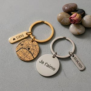 Op de afbeelding: Twee metalen sleutelhangers. Een goudkleurige sleutelhanger heeft een rechthoekig label met de inscriptie PARIS en een hartje, plus een ronde schijf met een zwarte stadsplattegrond. De zilverkleurige sleutelhanger heeft een schijf met de tekst Je t'aime en een label met de datum 03.04.2024.