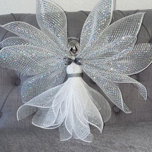 Op de afbeelding: Een decoratief engelenbeeldje met grote, glinsterende zilveren vleugels en een witte jurk. De engel heeft een zilveren halo en een kleine strik. De vleugels en jurk zijn gemaakt van een gaasmateriaal. De engel is een feestelijke decoratie.