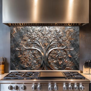 May include: A stainless steel cooker hood above a gas hob with a decorative metal splashback. The splashback features a detailed tree design with leaves and branches in silver and bronze. The kitchen appliances are set against a dark grey wall.