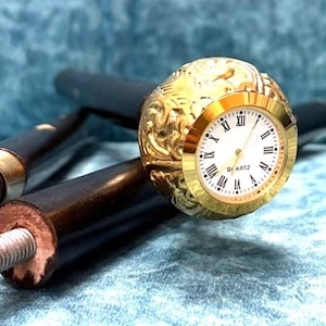 May include: A black walking cane with a gold-coloured clock face. The clock has Roman numerals and the word "Quartz" printed on the face. The cane has a gold-coloured metal accent.