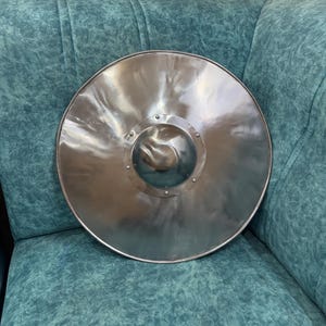 May include: A circular metal shield with a polished silver finish. The shield features a central domed boss, which is a raised hemispherical protrusion, secured to the main body by a series of small, evenly spaced metal rivets around its perimeter.