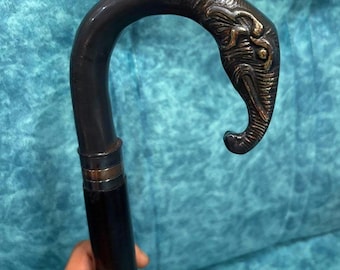 May include: A walking cane featuring a dark, curved handle shaped like an elephant head. The elephant head is crafted with metallic bronze-toned detailing on the eyes and trunk. The shaft of the cane is smooth, dark, and cylindrical, connected to the handle by a metallic band.